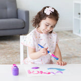 A young girl with a pink bow paints at a white table in a Bumkins Short-Sleeved Smock: Princess Magic, with a purple paint jar nearby.