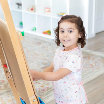 A young girl smiles at an easel, wearing a Bumkins Short-Sleeved Smock: Princess Magic in a bright room.