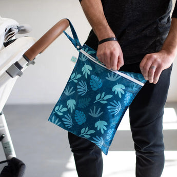 Someone attaches a Bumkins Blue Tropic wet bag, featuring a leaf pattern and waterproof fabric, to a stroller handle.