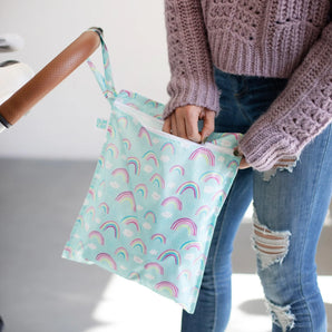 A person in a pink sweater and ripped jeans unzips a Bumkins Wet Bag: Rainbows hanging from a stroller handle.