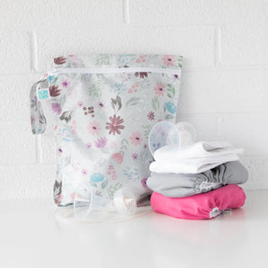 A Bumkins Wet Bag 2 Pack: Floral and Lace sits on a white table against a white brick wall with cloth diapers and breast pump parts in front.