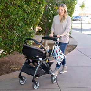 A smiling woman in jeans pushes a stroller with a Bumkins Wet Bag: Mickey Mouse Classic attached to the handle on a sidewalk past greenery.