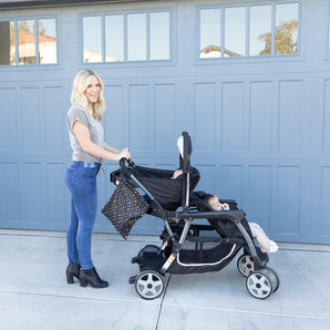 A person smiles with a child in a stroller by a blue garage, with Bumkins Wet Bag: Minnie Mouse Icon Black + White hanging from the handle.