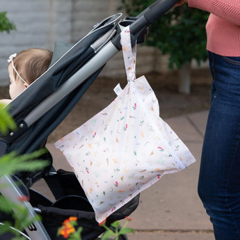 A person in jeans pushes a baby in a stroller outdoors, with Bumkins Wet Bag: Princess Magic hanging from the handle.