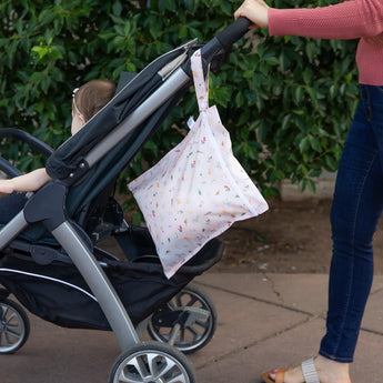 A person strolls with a baby in a stroller and Bumkins Wet Bag: Princess Magic hanging from the handle along a sidewalk lined with greenery.