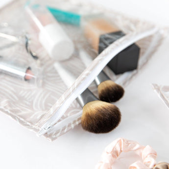Close-up of a Bumkins Clear Travel Bag 3-Pack: Wander with brushes, cosmetics and a scrunchie on a white background.
