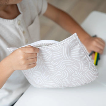 Close-up of a child at a white table placing colorful markers into a Bumkins Clear Travel Bag 3-Pack: Wander.
