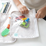 Close-up of a child's hands zipping a Bumkins Clear Travel Bag 3-Pack: Wander packed with toy cars and small items, resting on a white table.