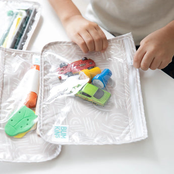 Close-up of a child's hands zipping a Bumkins Clear Travel Bag 3-Pack: Wander packed with toy cars and small items, resting on a white table.