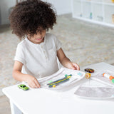 A child with curly hair plays with toys and art supplies in Bumkins Clear Travel Bag 3-Pack: Wander on a white table in a bright room.