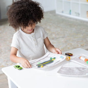 A child with curly hair plays with toys and art supplies in Bumkins Clear Travel Bag 3-Pack: Wander on a white table in a bright room.