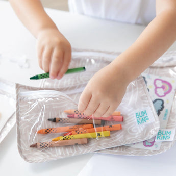 Close-up of a child placing a green crayon in a Bumkins Clear Travel Bag 3-Pack: Wander at a white table.