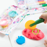 A child plays with a yellow rolling cutter on pink play dough near apple cutters, using Bumkins Clear Travel Bag 3-Pack for markers and dough.