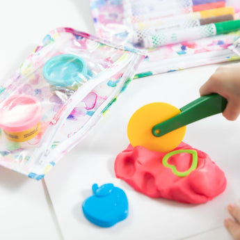A child plays with a yellow rolling cutter on pink play dough near apple cutters, using Bumkins Clear Travel Bag 3-Pack for markers and dough.
