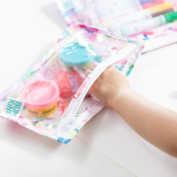 A child grabs playdough and markers from a Bumkins Clear Travel Bag, made from waterproof fabric, for endless fun.