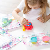 A young child with light brown hair is seated at a table playing with colorful playdough. Various art materials, including markers and TSA-compliant packages of playdough, are spread on the table in Bumkins Clear Travel Bag 3-Pack: Watercolor. The child is focused on her activity.