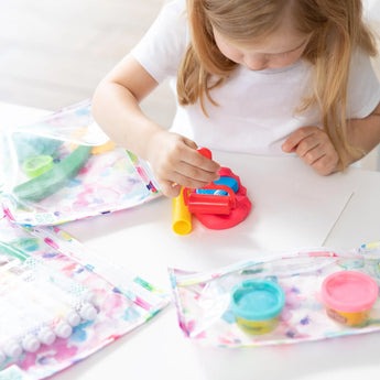 A young child with light brown hair is seated at a table playing with colorful playdough. Various art materials, including markers and TSA-compliant packages of playdough, are spread on the table in Bumkins Clear Travel Bag 3-Pack: Watercolor. The child is focused on her activity.