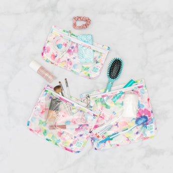A set of three TSA-friendly, watercolor floral Bumkins bags filled with beauty items sits on a marble surface.