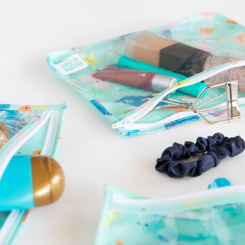 The Bumkins Clear Travel Bag 3-Pack: Ocean Life is beautifully displayed with makeup items, hair ties, and accessories on a white surface.
