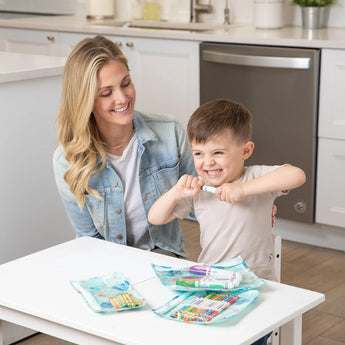 A woman and child sit with art supplies; the happy child holds a paintbrush, ready to design Bumkins Ocean Life reusable bags.