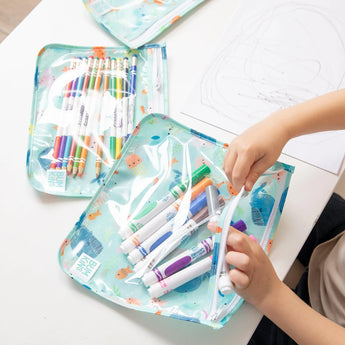 A child organizes colorful markers in a Bumkins ocean-themed pouch from the Clear Travel Bag 3-Pack, with a drawing sheet nearby.