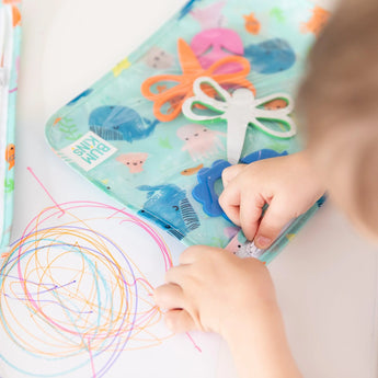 A child draws colorful doodles, surrounded by craft supplies in a Bumkins Clear Travel Bag 3-Pack: Ocean Life.
