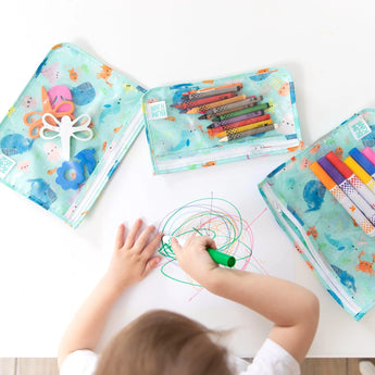 A child draws colorful scribbles with a green marker on white paper. Nearby, three Bumkins Clear Travel Bags featuring ocean life patterns hold scissors and various markers. The scene unfolds on a white surface.