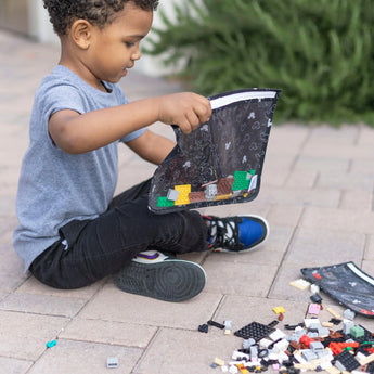 A child pours colorful blocks from a Bumkins Clear Travel Bag: Mickey Mouse Icon Black + White onto the pavement.