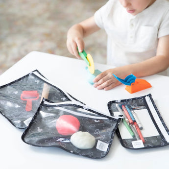 A child plays with modeling clay at a table, surrounded by Bumkins Clear Travel Bag 3-Pack: Mickey Mouse Icon Black + White. These pouches feature a black pattern and white zippers, containing extra clay, tools, and pens against a softly blended background.