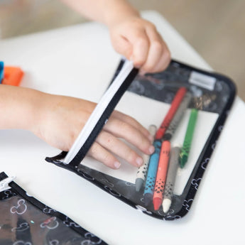A child opens a Bumkins Mickey Mouse black & white clear travel bag full of colorful pens and pencils on a white table.