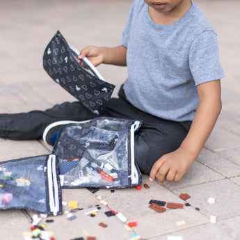 A child plays with toy bricks next to Bumkins TSA-compliant Mickey Mouse Icon Black + White Clear Travel Bag 3-Pack filled with more bricks.